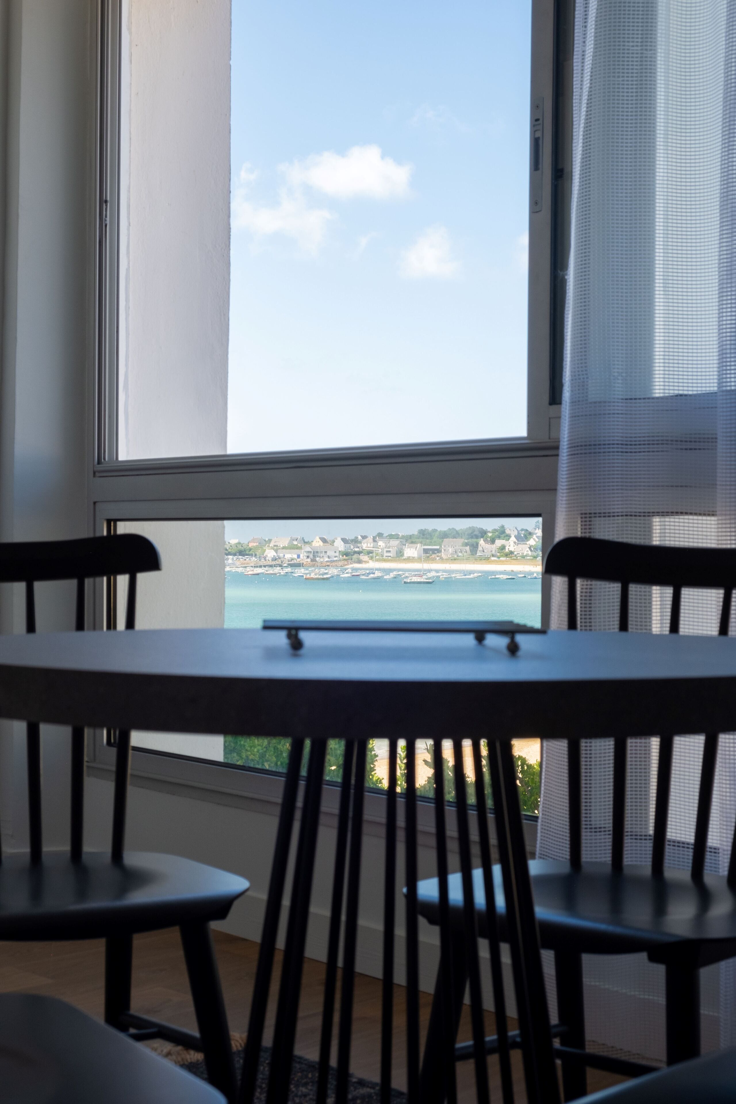 A room with a table and chairs facing a large window overlooking a coastal view.