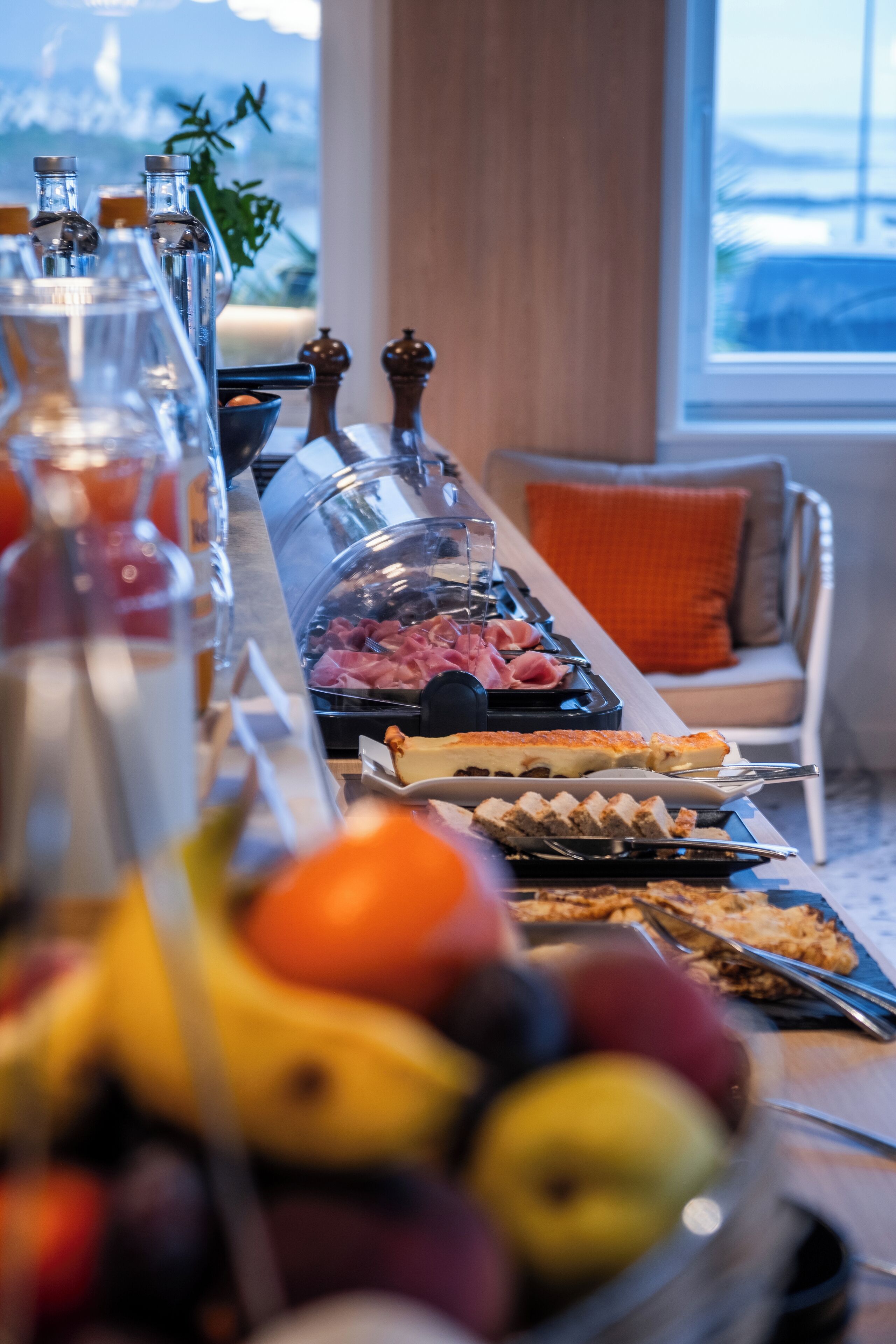 Un bufé de desayuno con diversas opciones de comida y bebidas, una ventana con vistas y un rincón de descanso acogedor con cojín.