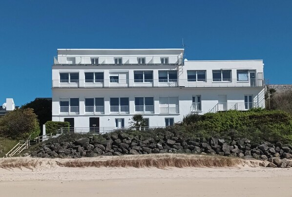Een meerlaags wit gebouw met balkons, nabij een zandstrand met een rotsachtige barrière.