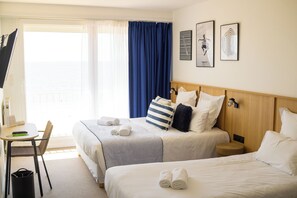 Een hotelkamer met twee bedden, een bureau en een stoel. Er liggen handdoeken op de bedden en er is een raam met gordijnen.