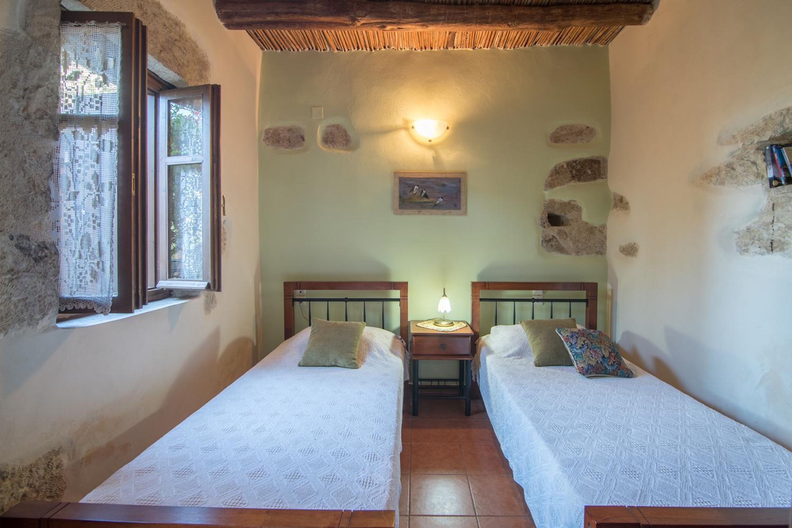 A small, rustic room with two single beds, a nightstand with a lamp, and a window with lace curtains.