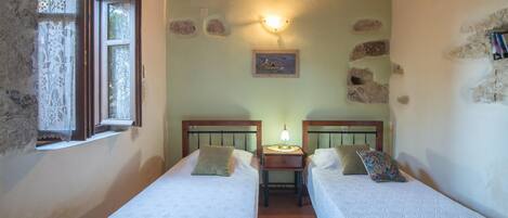 Une petite chambre rustique avec deux lits simples, une table de chevet avec une lampe et une fenêtre ornée de rideaux en dentelle.