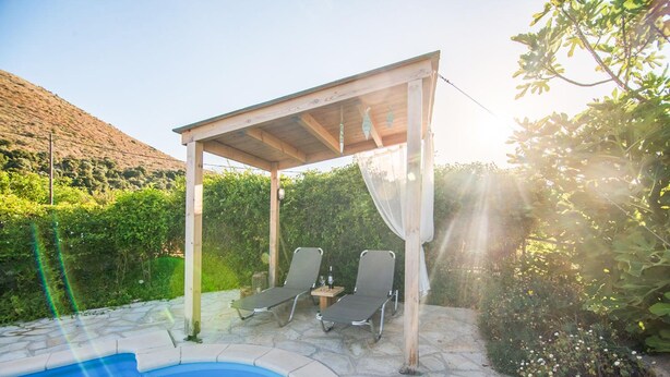 A pool area with two lounge chairs under a pergola, surrounded by stone tiles and greenery. - Villa Kourkoulios in Apokoronos (Apokoronas)