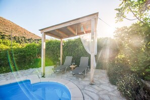 A pool area with two lounge chairs under a pergola, surrounded by stone tiles and greenery. - Villa Kourkoulios in Apokoronos (Apokoronas)
