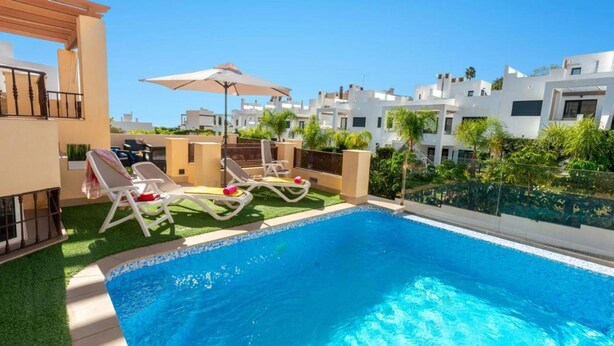 A swimming pool with lounge chairs and an umbrella, surrounded by greenery and modern buildings. - Casa Eva in Nerja With 3 Bedrooms and 3 Bathrooms (Nerja)