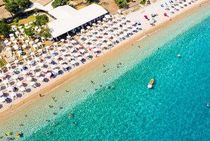 An aerial view of a beach with sun loungers and umbrellas, a clear blue sea, and a building with a white roof. - Villa Emily in Apokoronos (Corfu)