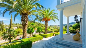 A well-maintained garden with palm trees, neatly trimmed hedges, and a stone staircase leading to a covered area with columns.