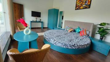 A modern bedroom with a round bed, a blue round table, a wicker chair, and a television on a stand.