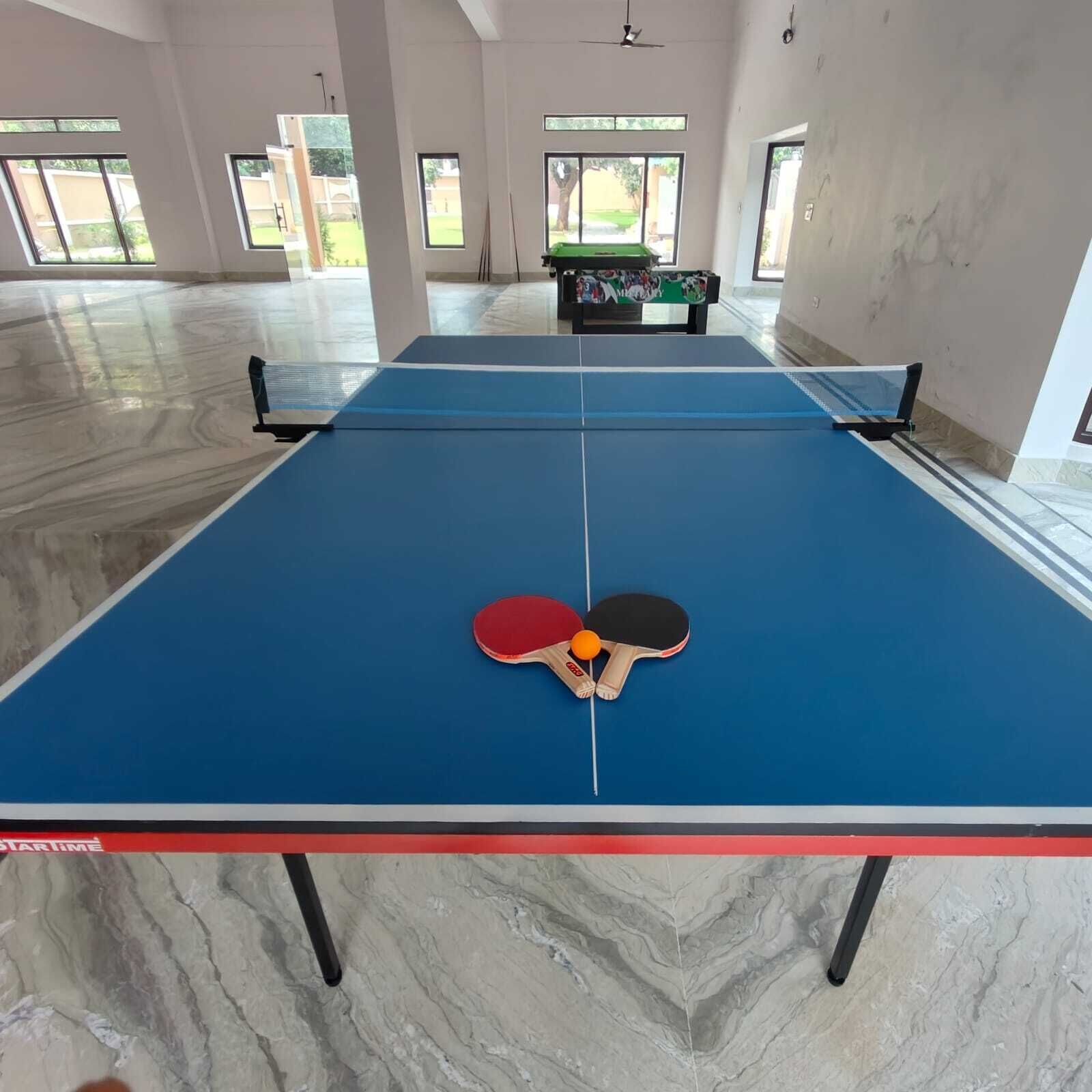 Una mesa de ping pong con dos paletas y una pelota en el centro, rodeada por una habitación amplia con ventanas grandes y una mesa de billar al fondo.