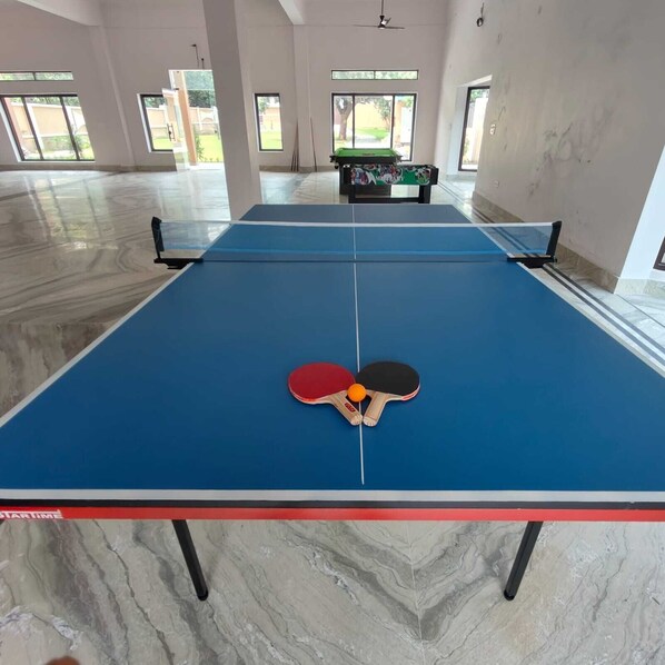 A ping pong table with two paddles and a ball in the center, surrounded by a spacious room with large windows and a pool table in the background. - Atithi Stays (Ramnagar)