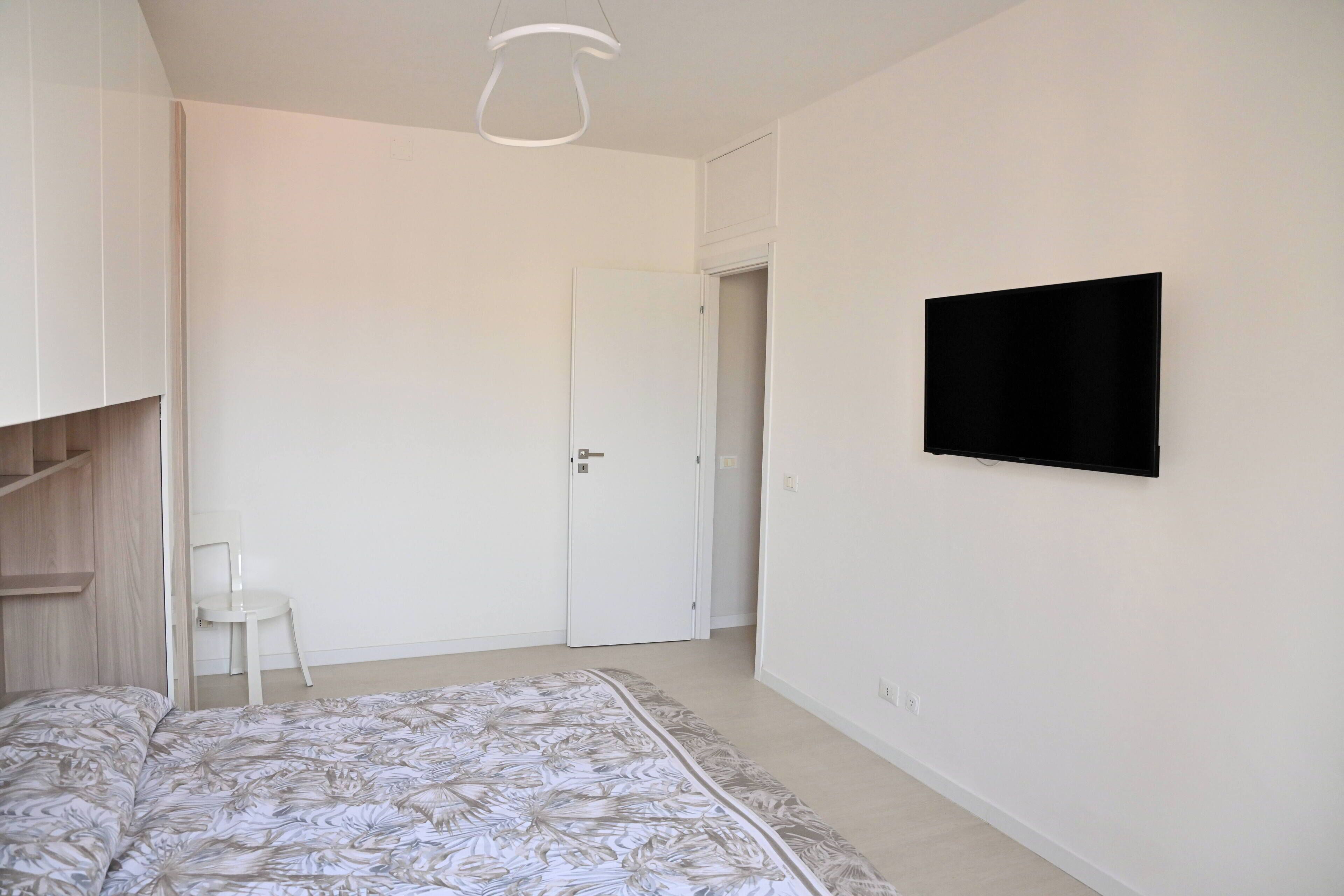 Une chambre à coucher comprenant un lit, un téléviseur fixé au mur, une chaise et une porte donnant sur une autre pièce.