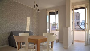 A dining area with a wooden table and white chairs, a grey sofa, and a patterned wallpaper. - Emerenziana Lovely Apartment (Rome)