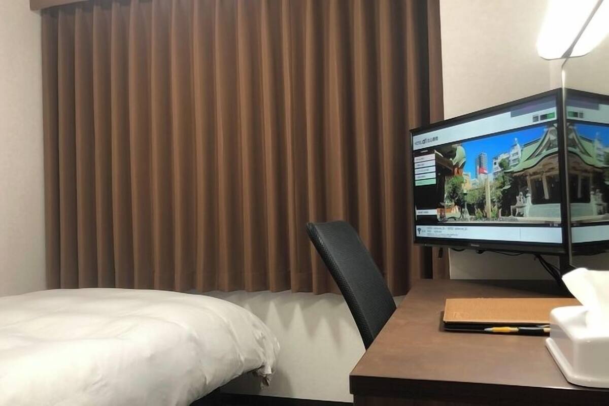 Une chambre d’hôtel avec un lit, un bureau doté d’un écran d’ordinateur et une fenêtre avec des rideaux marron.