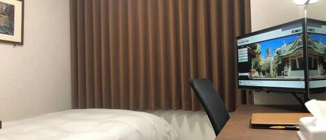 Ein Hotelzimmer mit einem Bett, einem Schreibtisch mit Computerbildschirm und einem Fenster mit braunen Vorhängen.