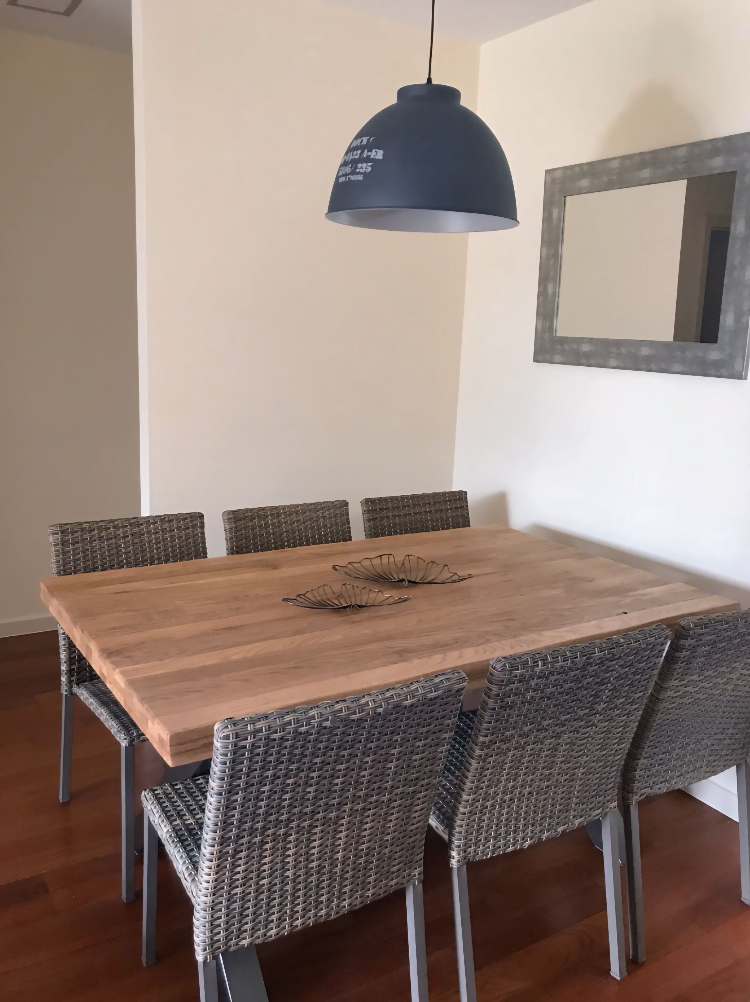 Un comedor con mesa de madera y sillas de mimbre, una lámpara colgante oscura y un espejo en la pared.