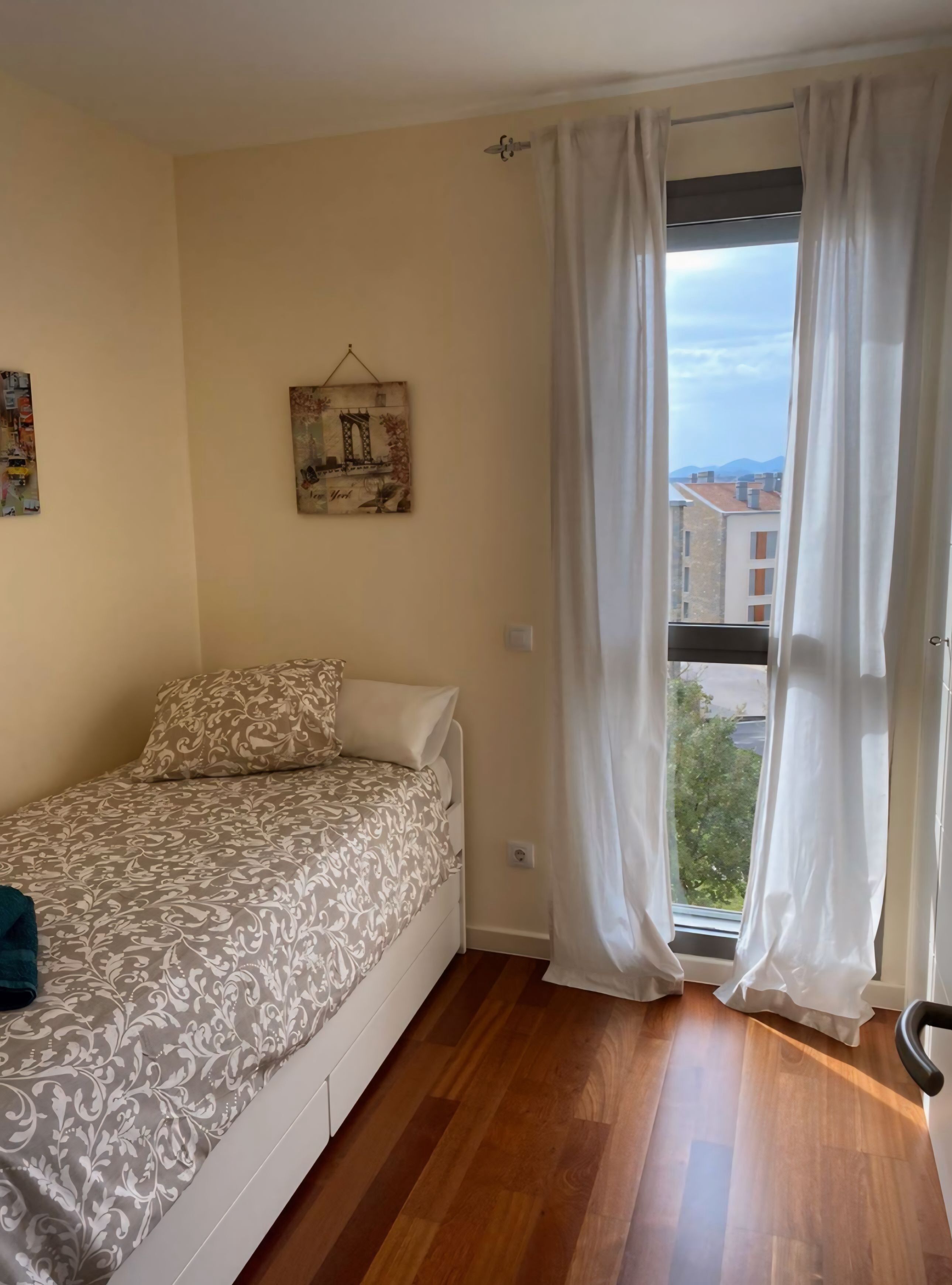 Un dormitorio pequeño con cama, ventana con cortinas, un cuadro en la pared y vistas a edificios y árboles.