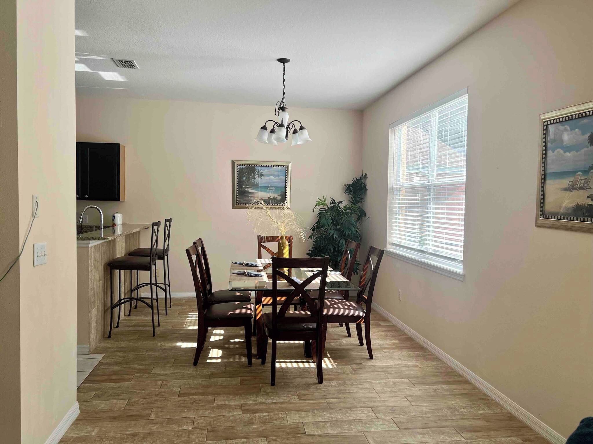 A dining area with a chandelier, wooden chairs, a glass table, and a painting on the wall.