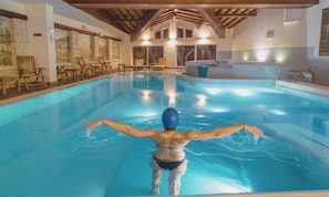 An indoor swimming pool with a person swimming, wooden chairs, and a sauna area.