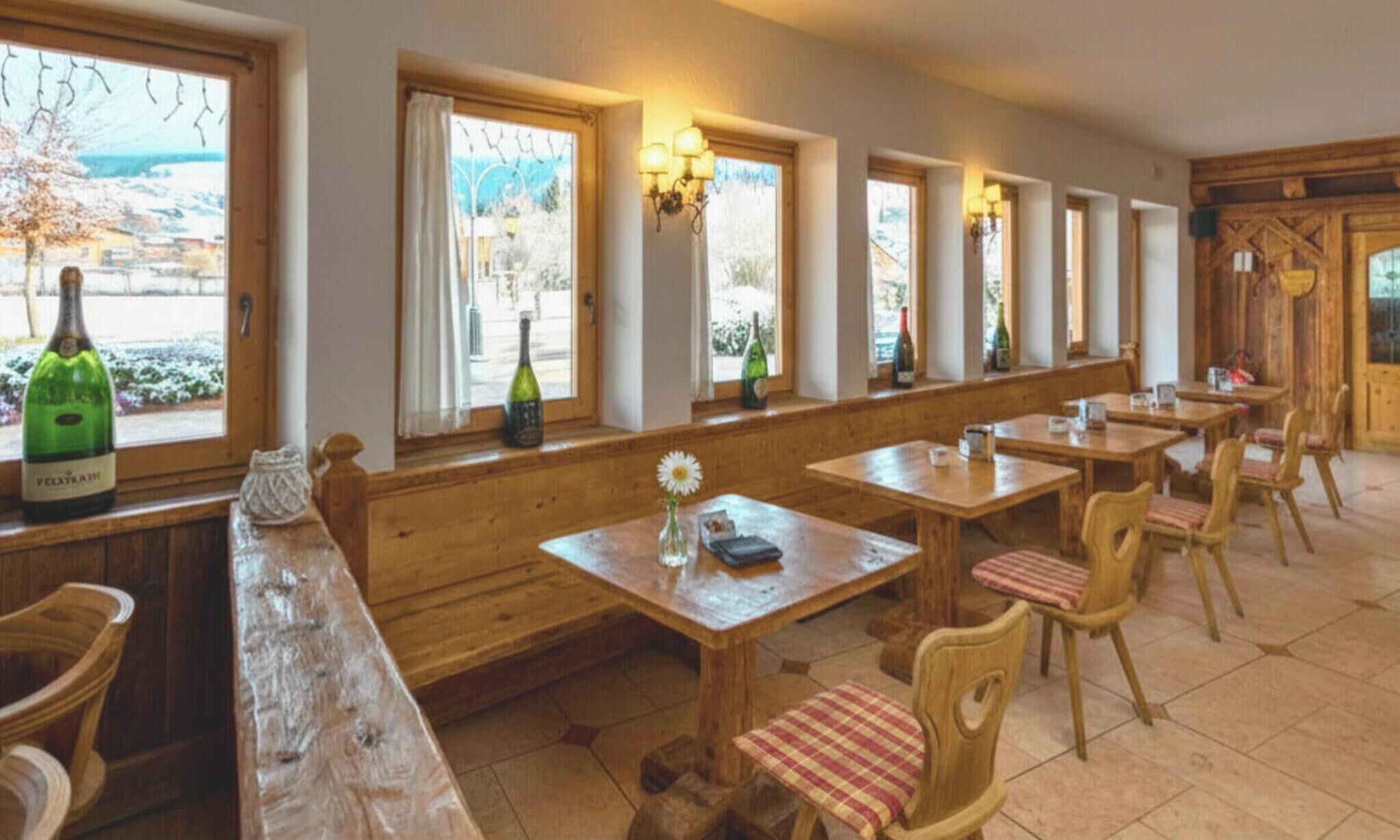 A restaurant with wooden tables and chairs, a bench, and bottles of wine on the windowsill.