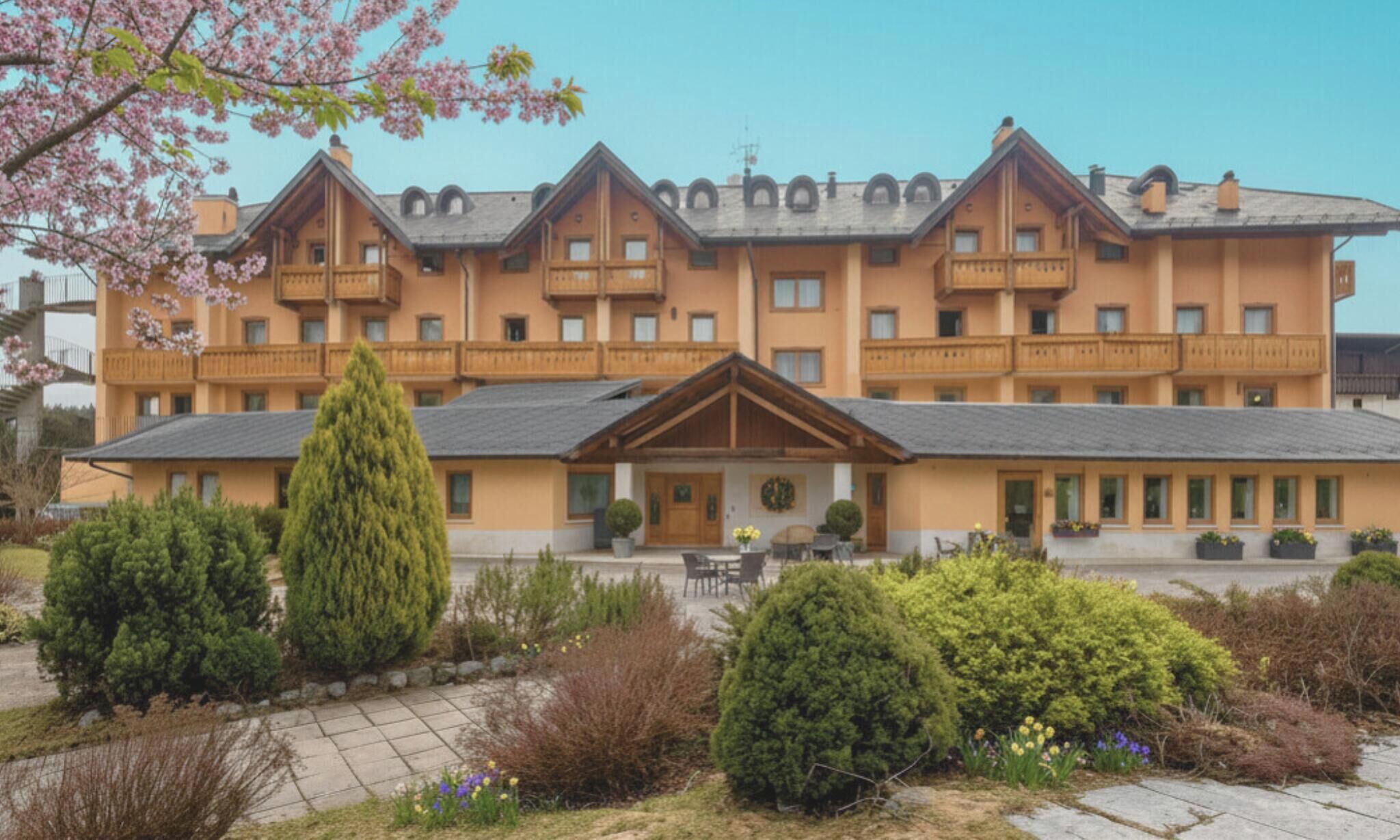 A multi-story hotel with balconies, a central entrance, and a garden area with shrubs and flowers.