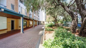 A corridor with a brick floor, a covered walkway, and a row of rooms with windows and doors.