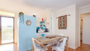 A dining area with a rustic wooden table and white chairs, a blue door, and a kitchen in the background.