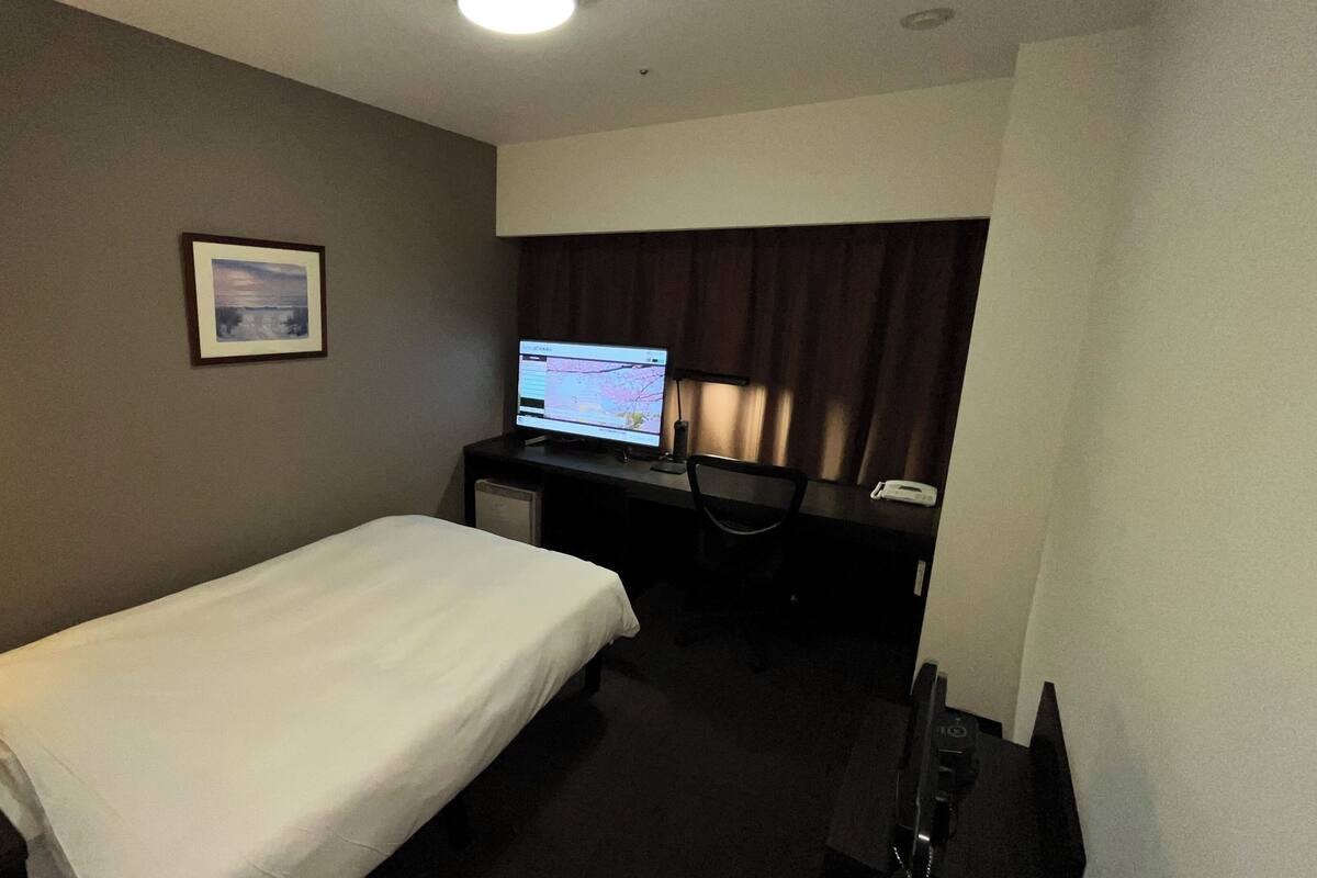 A hotel room with a single bed, a desk with a computer, a telephone, and a framed picture on the wall.