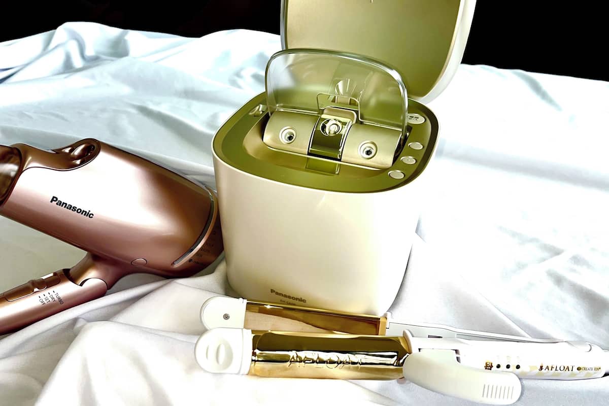 A Panasonic hairdryer, a white and gold hairbrush, and a white and gold hair straightener on a white fabric surface.