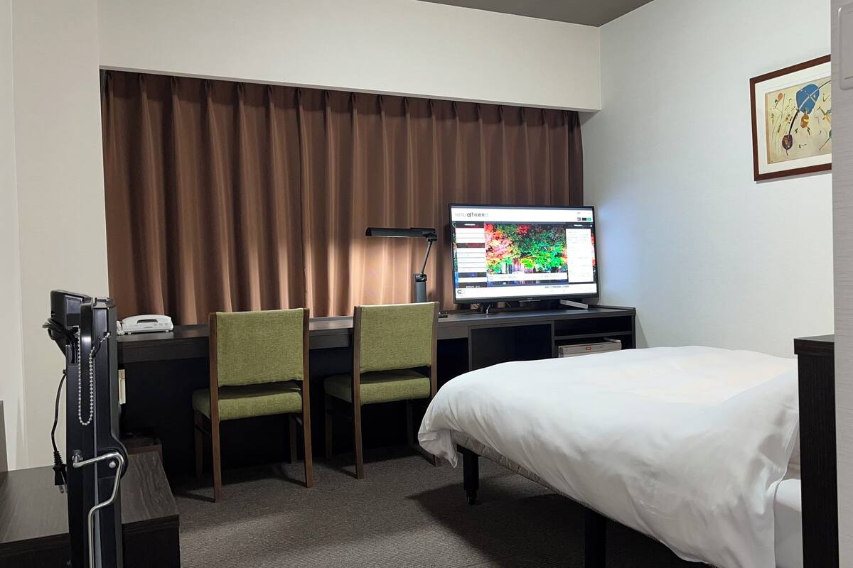 A hotel room with a bed, a desk with a computer, two chairs, a telephone, and a window with curtains.