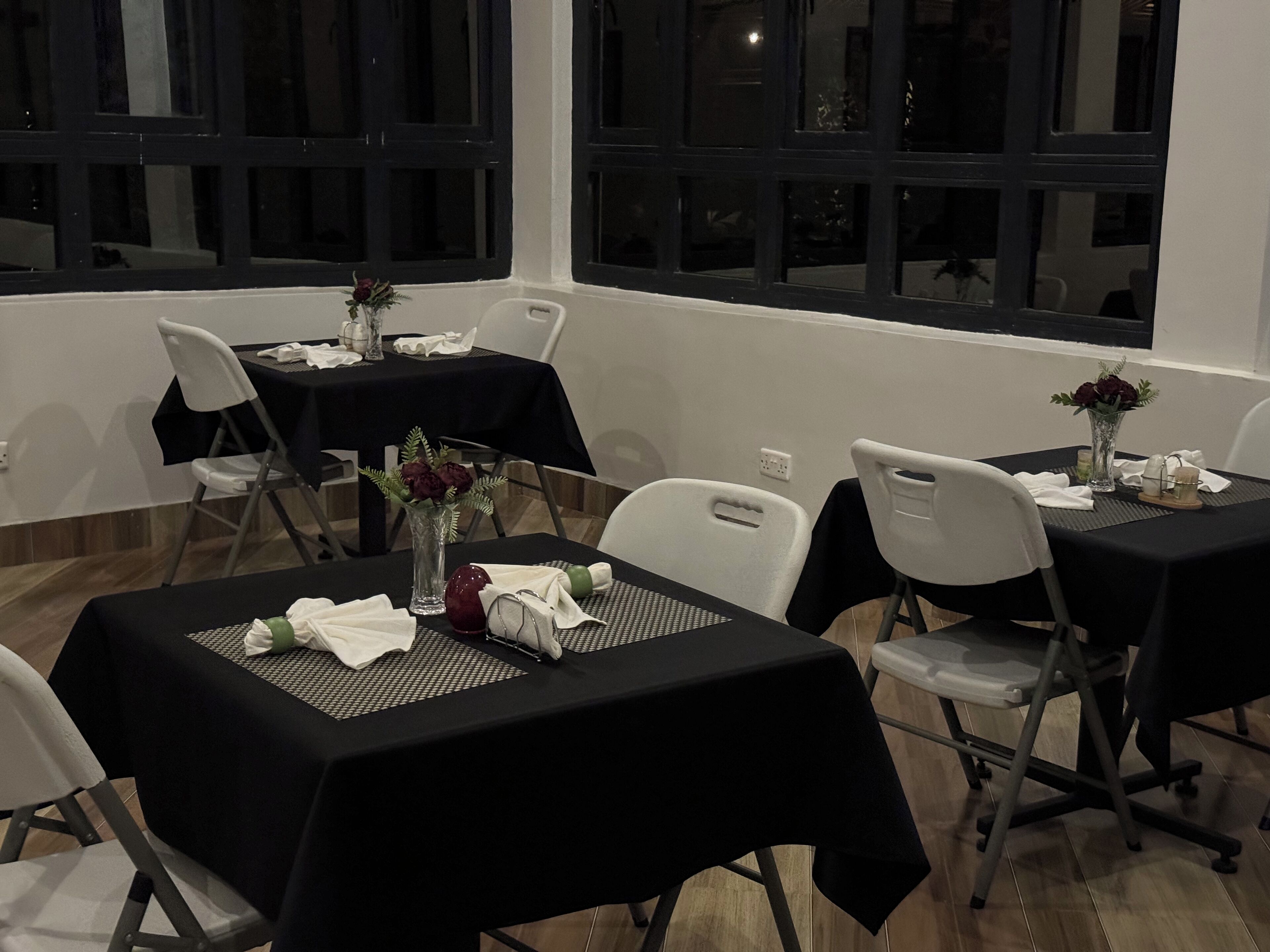 A dining area with tables set for four, each with a black tablecloth, white chairs, and a vase with flowers.