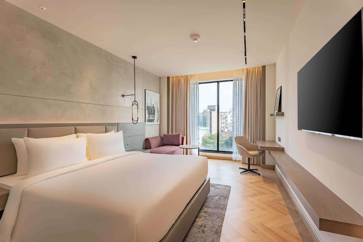 A modern hotel room with a large bed, a flat-screen TV, a seating area, and a view of the city through large windows.