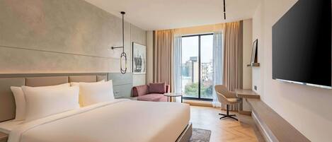 Una habitación de hotel moderna con una cama grande, un televisor de pantalla plana, un área de estar y vistas a la ciudad a través de amplios ventanales.