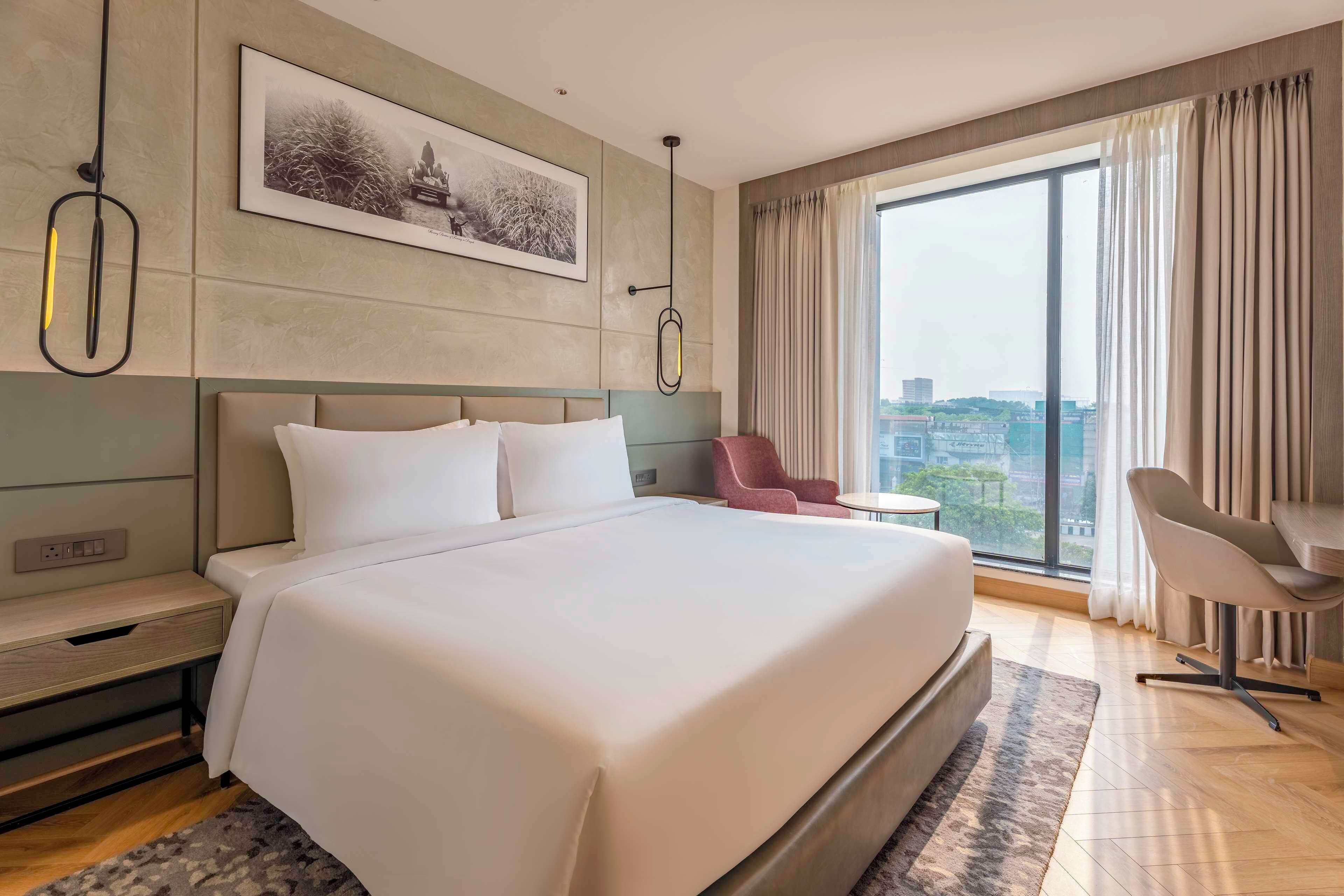 A modern hotel room with a large bed, a desk, a chair, and a view of the city through large windows.