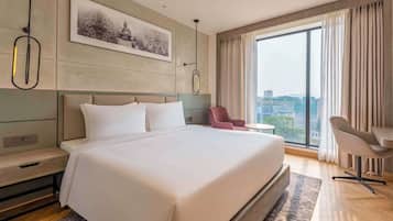 A modern hotel room with a large bed, a desk, a chair, and a view of the city through large windows.
