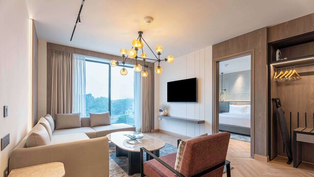 A modern hotel room with a sofa, armchair, coffee table, and a TV mounted on the wall. There is a large window with curtains, a wooden floor, and a built-in closet with a mirror and hanging clothes. - Park Inn By Radisson Jalandhar (Jalandhar)