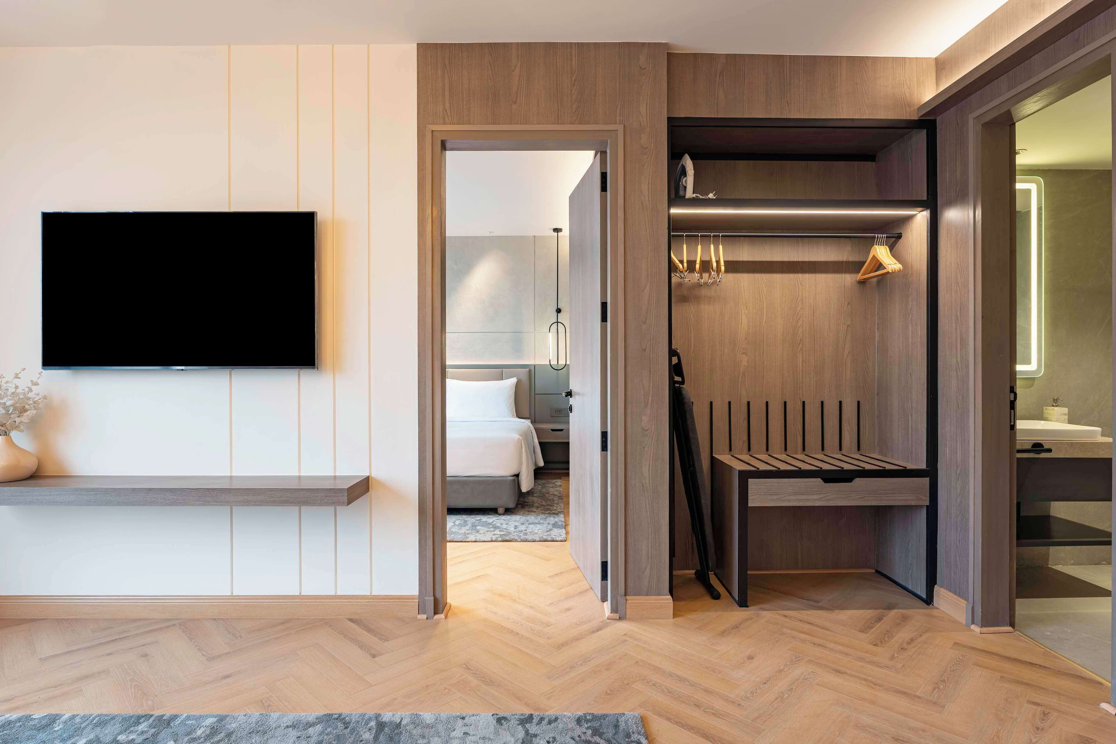 A modern hotel room with a flat-screen TV mounted on the wall, a wooden bench, a walk-in closet with a mirror, and a visible bathroom.