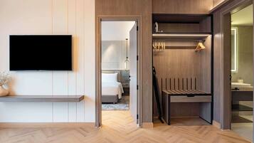 A modern hotel room with a flat-screen TV mounted on the wall, a wooden bench, a walk-in closet with a mirror, and a visible bathroom.