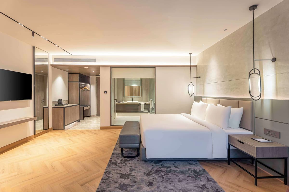 A modern hotel room with a large bed, a desk, a bench, a TV, and a walk-in closet.