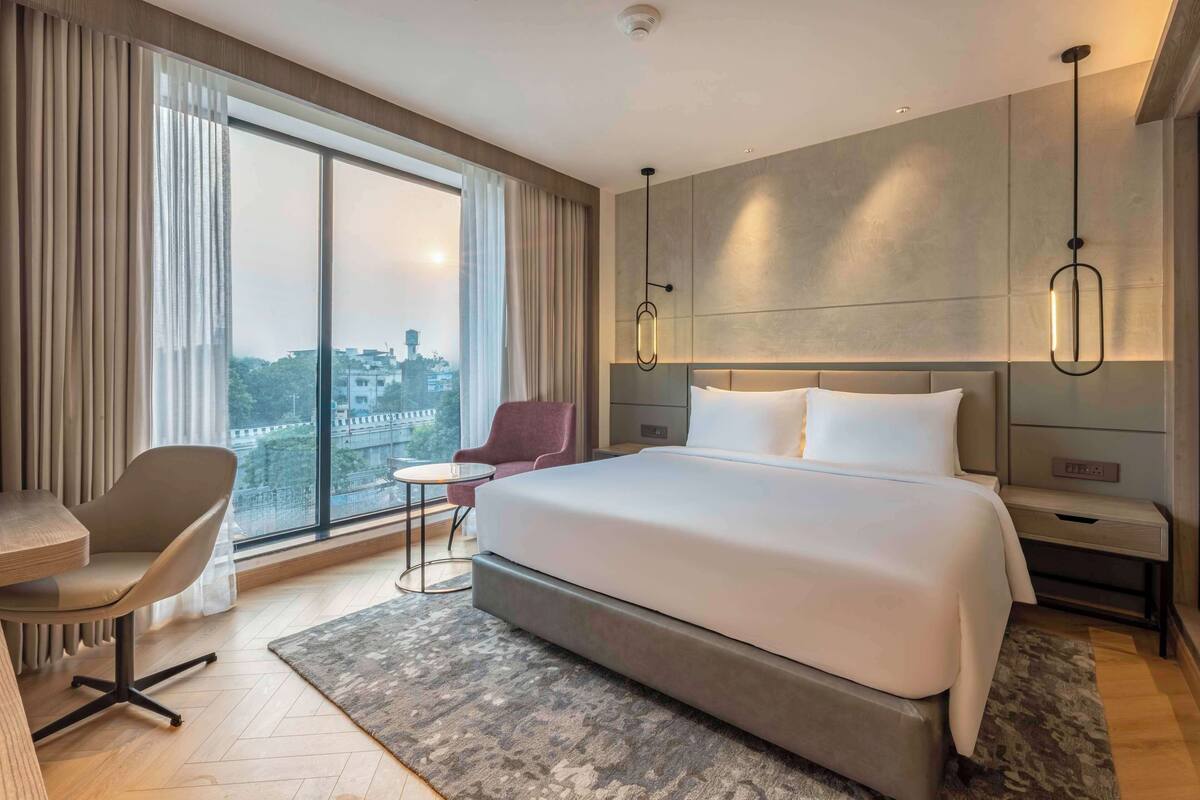 A modern hotel room with a large bed, a desk, a chair, and a view of the city through large windows.