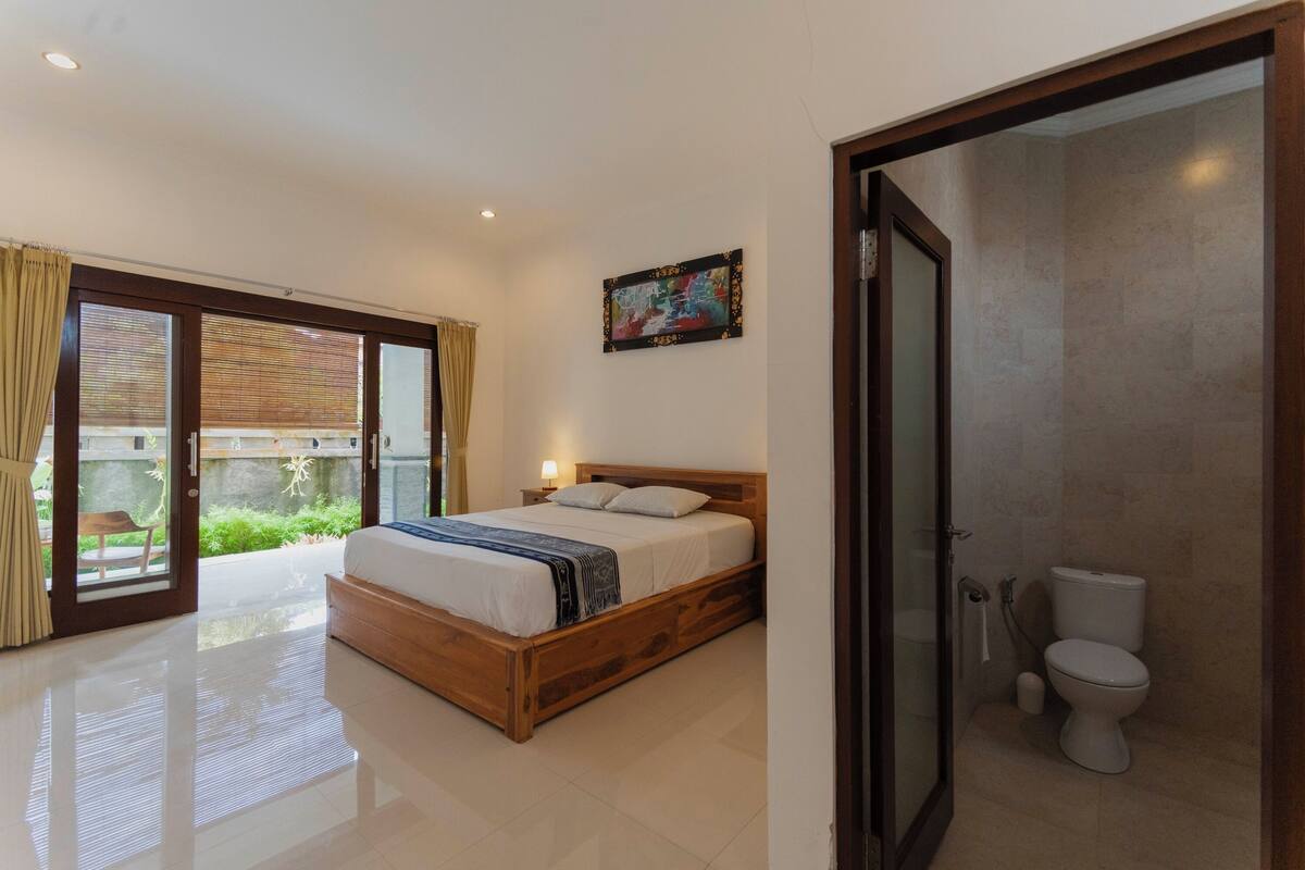 Un dormitorio con cama de madera, un baño con inodoro y un ventanal corredero que da a un balcón.