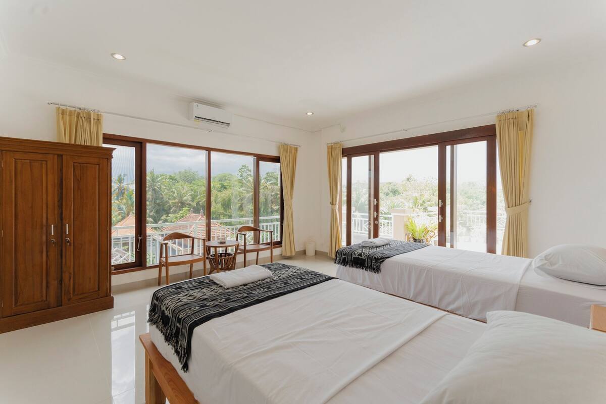 Habitación de hotel con dos camas, un armario de madera, una mesita con una silla y grandes ventanales que ofrecen vistas a zonas verdes y edificios.