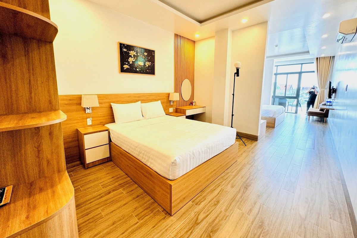 A modern hotel room with a large bed, wooden headboard, bedside tables, a nightstand, a floor lamp, a painting on the wall, and a view of the outdoors through the window.