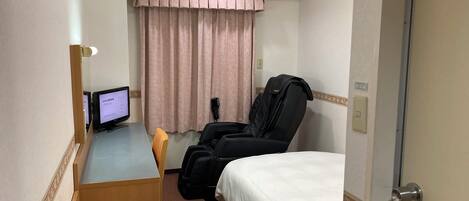 A hotel room with a bed, a massage chair, a desk with a computer, and a window with curtains.
