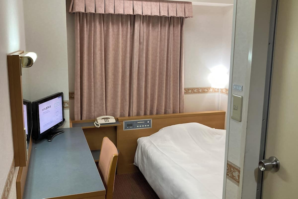 Ein Hotelzimmer mit einem Bett, einem Schreibtisch mit Computer, einem Telefon und einem Fenster mit Vorhängen.