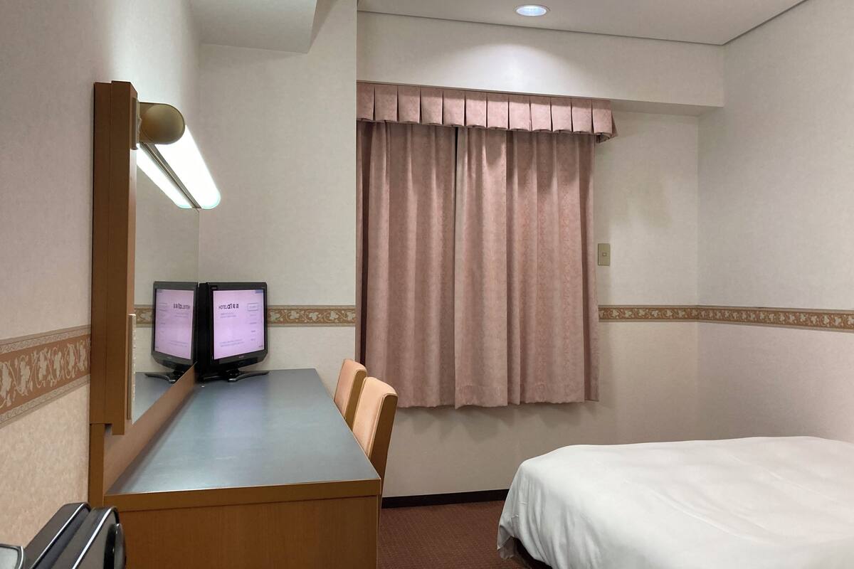 Ein Hotelzimmer mit Schreibtisch, zwei Bildschirmen, einem Bett, einem Fenster mit Vorhängen und einem gerahmten Bild an der Wand.