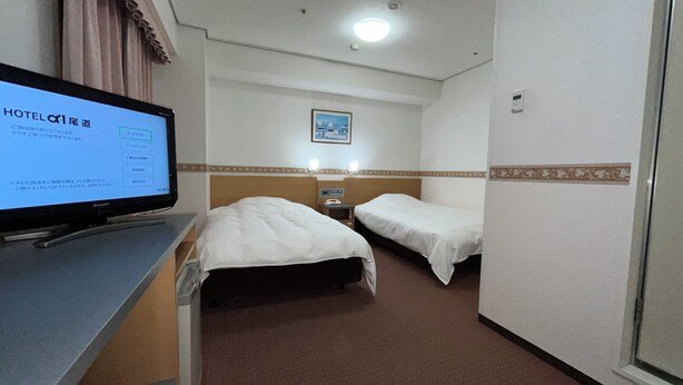 A hotel room with two beds, a desk with a computer, and a wall-mounted TV displaying a hotel directory. - Hotel Alpha-One Onomichi (Onomichi)