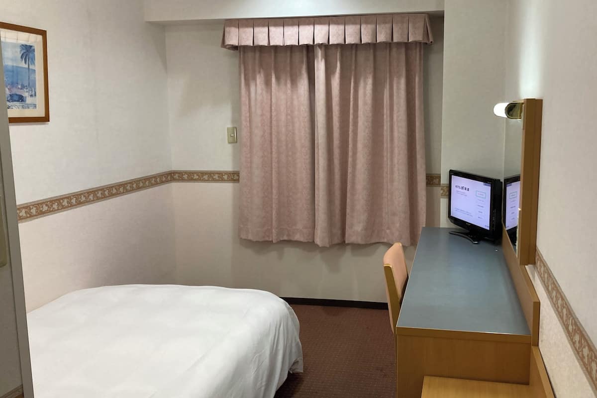 Ein Hotelzimmer mit einem Bett, einem Schreibtisch mit Computer, einem Stuhl, einem Fenster mit Vorhängen und einem gerahmten Bild an der Wand.