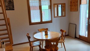 A room with a round wooden table and four chairs, a wooden ladder, a window with lace curtains, a mirror, and a radiator.