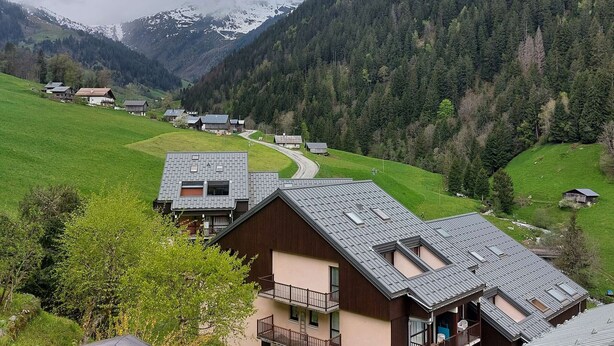 A rural mountain village with traditional wooden houses, a winding road, and a backdrop of snow-capped mountains.