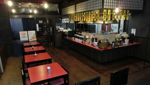 Interno di un ristorante con diversi tavoli rossi e sedie nere, un bancone con varie attrezzature da cucina e insegne del menu in giapponese.